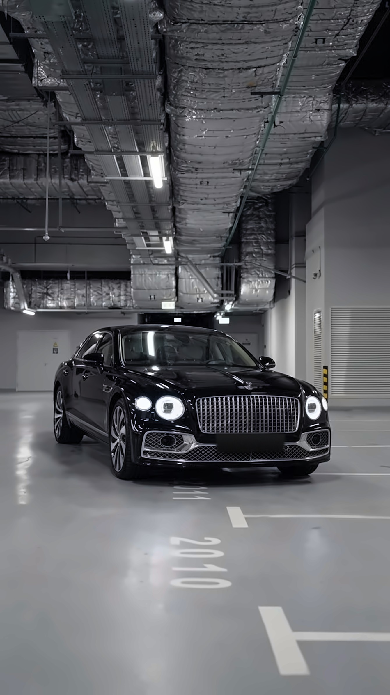 A black Bentley Flying Spur with its headlights on, freshly washed and gleaming, parked in spot 2010 in a residential garage at night